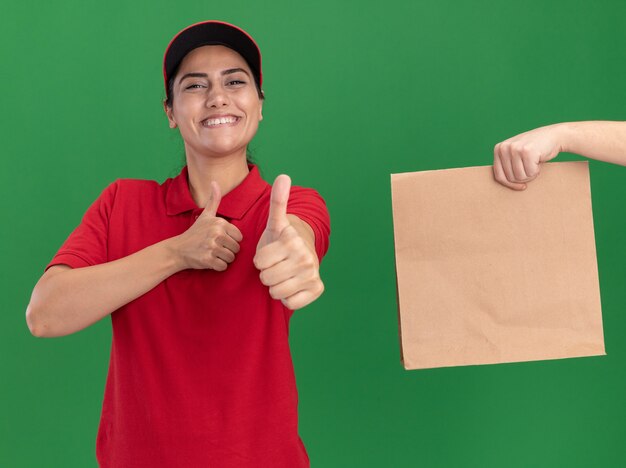 Smiling young delivery girl wearing uniform and cap showing thumb up someone giving money to her isolated on green wall
