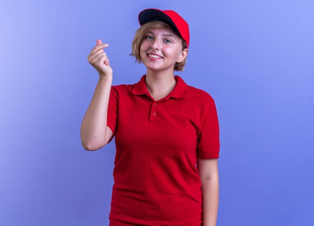 Smiling young delivery girl wearing uniform and cap showing tip gesture isolated on blue wall