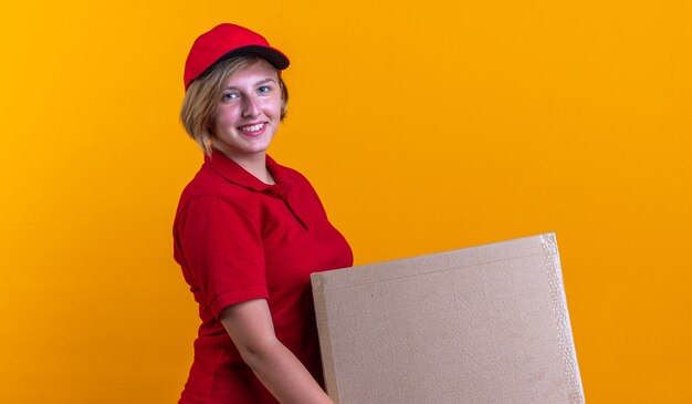 smiling young delivery girl wearing uniform with cap holding box isolated on orange wall