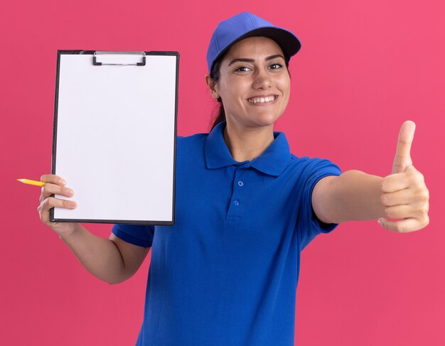 Smiling young delivery girl wearing uniform with cap holding clipboard and showing thumb up isolated on pink wall