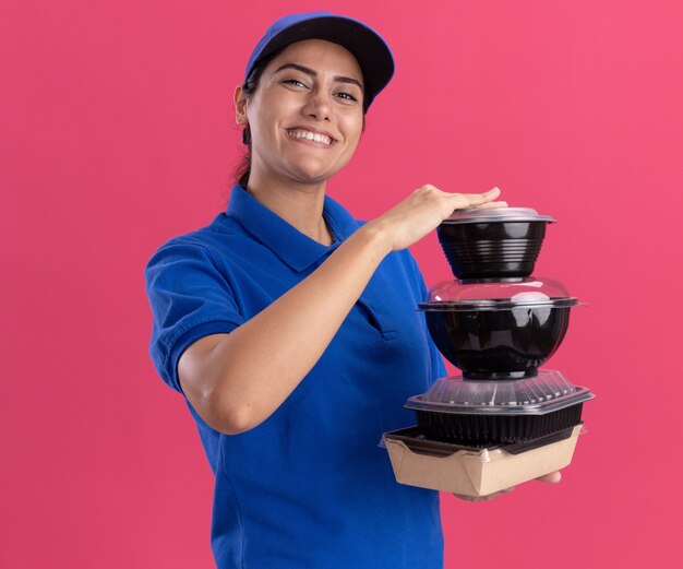 Smiling young delivery girl wearing uniform with cap holding food containers isolated on pink wall