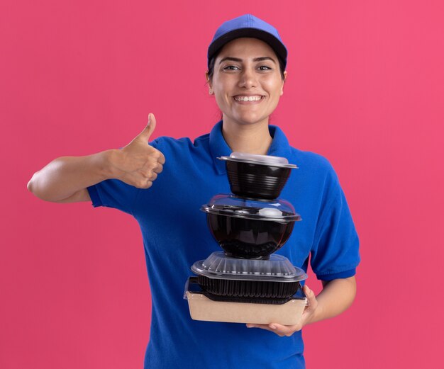 Smiling young delivery girl wearing uniform with cap holding food containers showing thumb up isolated on pink wall