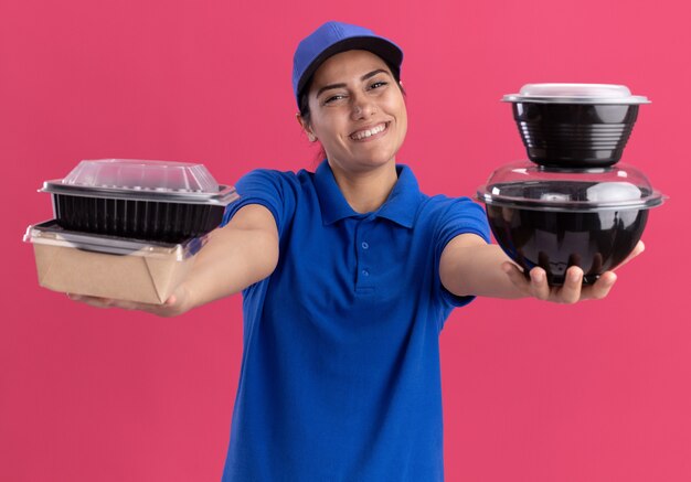 Smiling young delivery girl wearing uniform with cap holding out food containers at front isolated on pink wall