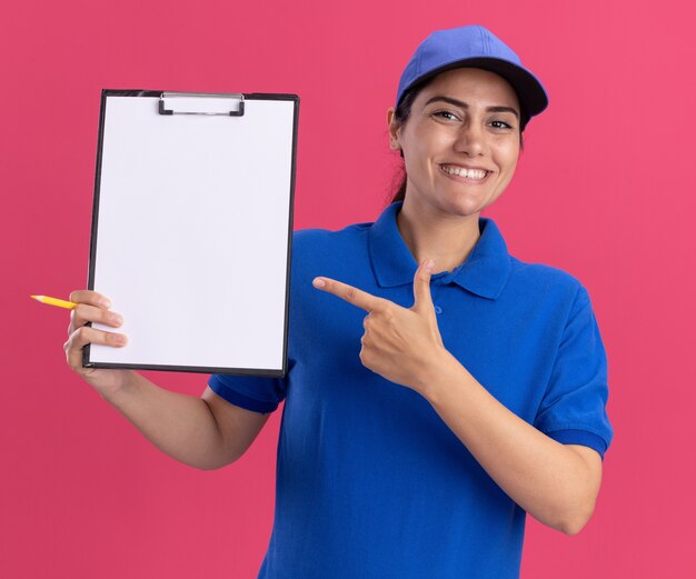 Smiling young delivery girl wearing uniform with cap holding and points at clipboard isolated on pink wall