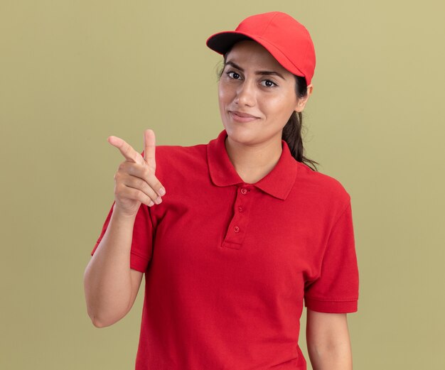 Smiling young delivery girl wearing uniform with cap points at camera isolated on olive green wall