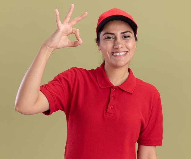 Улыбающаяся молодая доставщица в униформе с кепкой, показывающая нормальный жест, изолирована на оливково-зеленой стене