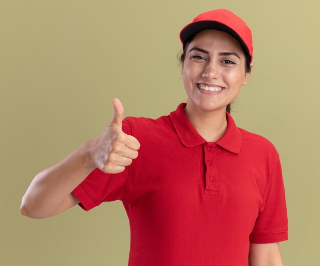 オリーブグリーンの壁に分離された親指を示すキャップと制服を着て笑顔の若い配達の女の子
