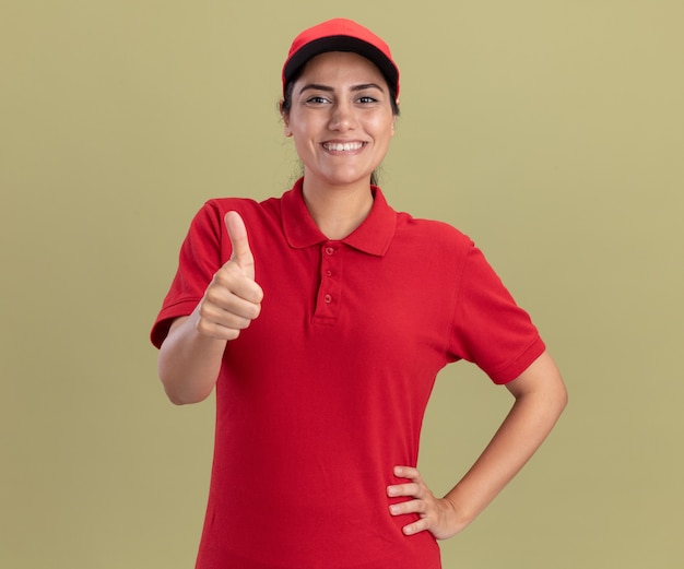 Sorridente giovane ragazza delle consegne che indossa l'uniforme con il cappuccio che mostra il pollice in su e mette la mano sull'anca isolata sulla parete verde oliva