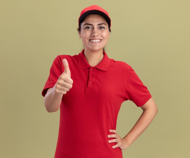 Smiling young delivery girl wearing uniform with cap showing thumb up and putting hand on hip isolated on olive green wall