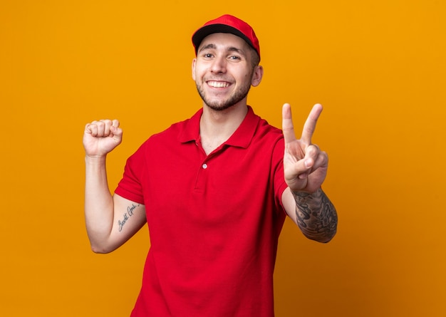 Smiling young delivery guy wearing uniform with cap showing peace gesture 