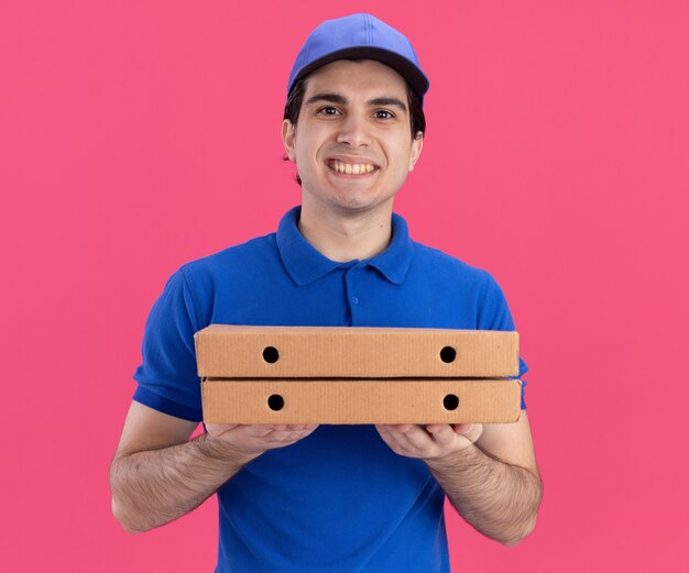 Sorridente giovane fattorino in uniforme blu e berretto che tiene i pacchetti di pizza guardando la parte anteriore isolata sulla parete rosa