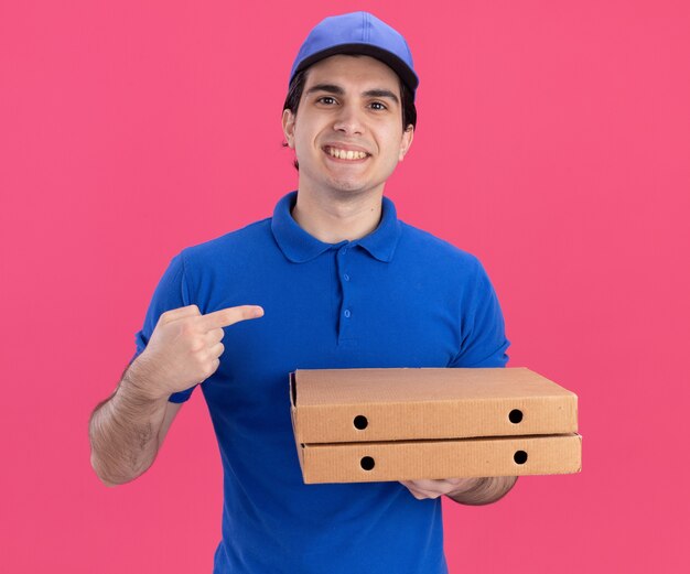 Smiling young delivery man in blue uniform and cap holding pizza packages looking at front pointing at side isolated on pink wall