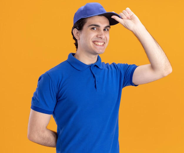 Smiling young delivery man in blue uniform and cap looking at front keeping hand behind back grabbing cap isolated on orange wall