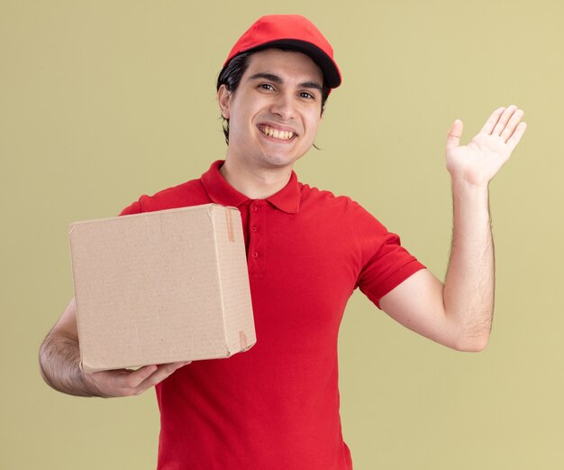 Smiling young delivery man in red uniform and cap holding cardbox looking at front showing empty hand isolated on olive green wall