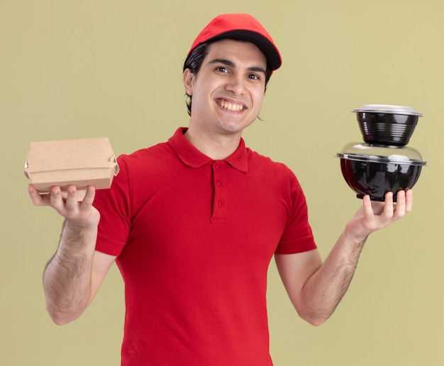 Smiling young delivery man in red uniform and cap holding paper food package and food containers looking at front isolated on olive green wall