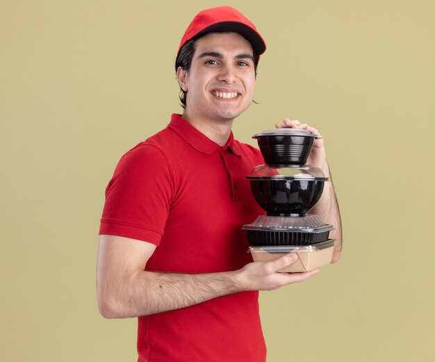 Smiling young delivery man in red uniform and cap standing in profile view holding food containers and paper food package looking at front isolated on olive green wall