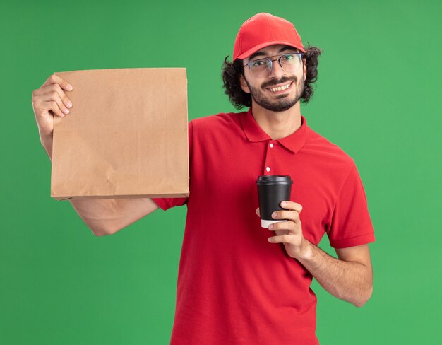 Smiling young delivery man in red uniform and cap wearing glasses holding paper package and plastic coffee cup looking at front isolated on green wall