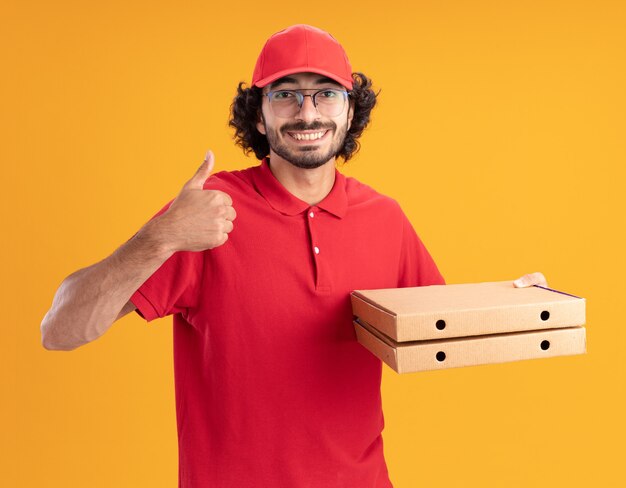 Smiling young delivery man in red uniform and cap wearing glasses holding pizza packages looking at front showing thumb up isolated on orange wall