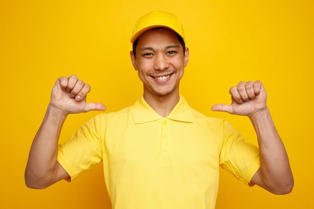 Smiling young delivery man wearing cap and uniform pointing at himself 