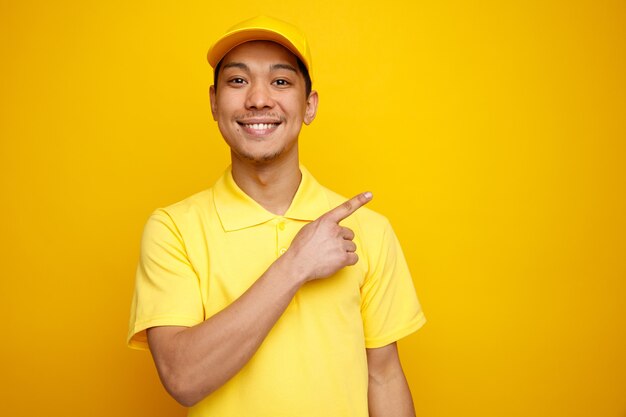 Smiling young delivery man wearing cap and uniform pointing up at corner 