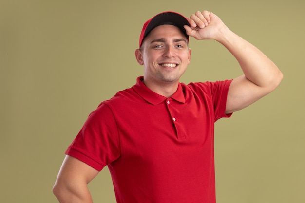 Smiling young delivery man wearing uniform and cap holding cap isolated on olive green wall