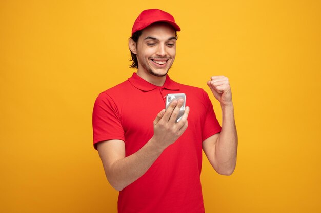 Улыбающийся молодой курьер в форме и кепке держит мобильный телефон и смотрит на него, показывая жест "да" на желтом фоне
