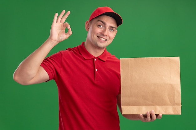 Smiling young delivery man wearing uniform and cap holding paper food package showing okay gesture isolated on green wall