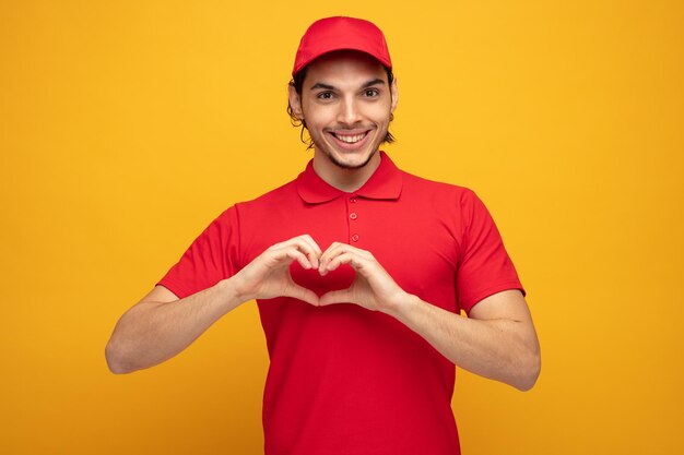 smiling young delivery man wearing uniform and cap looking at camera isolated on yellow background