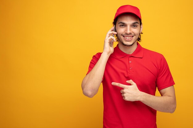 smiling young delivery man wearing uniform and cap looking at camera pointing to side isolated on yellow background with copy space