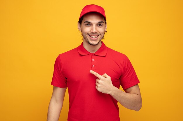 smiling young delivery man wearing uniform and cap looking at camera pointing to side isolated on yellow background