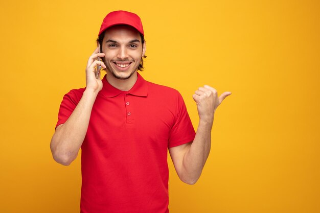 노란색 배경에 격리된 전화 통화 중 옆을 가리키는 카메라를 바라보며 유니폼을 입고 모자를 쓴 웃고 있는 젊은 배달원