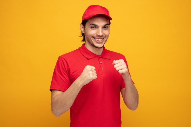 Улыбающийся молодой курьер в форме и кепке смотрит в камеру, показывая боксерский жест на желтом фоне