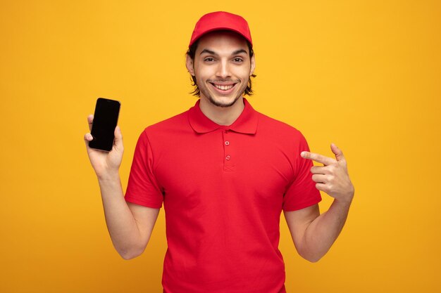 smiling young delivery man wearing uniform and cap looking at camera showing mobile phone pointing at it isolated on yellow background