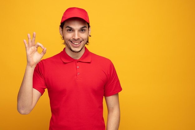 smiling young delivery man wearing uniform and cap looking at camera showing ok sign isolated on yellow background with copy space