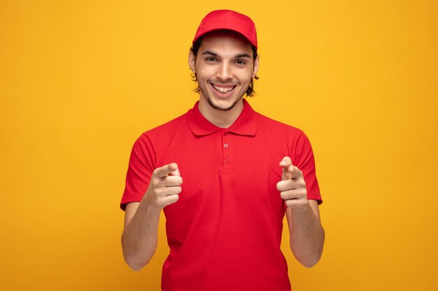 smiling young delivery man wearing uniform and cap looking at camera showing you gesture isolated on yellow background