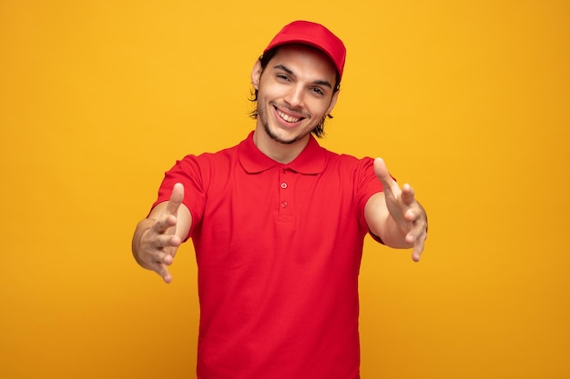 Sorridente giovane fattorino che indossa uniforme e berretto guardando la fotocamera allungando le mani verso la fotocamera isolata su sfondo giallo