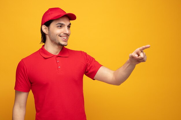 smiling young delivery man wearing uniform and cap looking and pointing at side isolated on yellow background