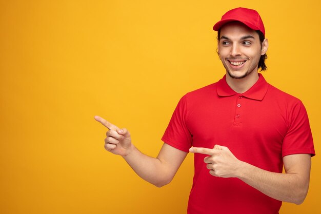 smiling young delivery man wearing uniform and cap looking at side pointing to side isolated on yellow background with copy space