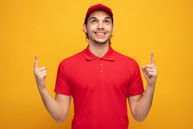 黄色の背景に分離された指を上に向けて見上げる制服とキャップを身に着けている若い配達人の笑顔