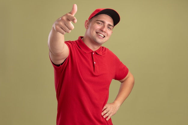 Sorridente giovane fattorino che indossa l'uniforme e berretto che mostra il gesto e mette la mano sull'anca isolata sulla parete verde oliva