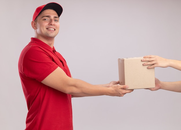 Smiling young delivery man wearing uniform with cap giving box to client isolated on white wall