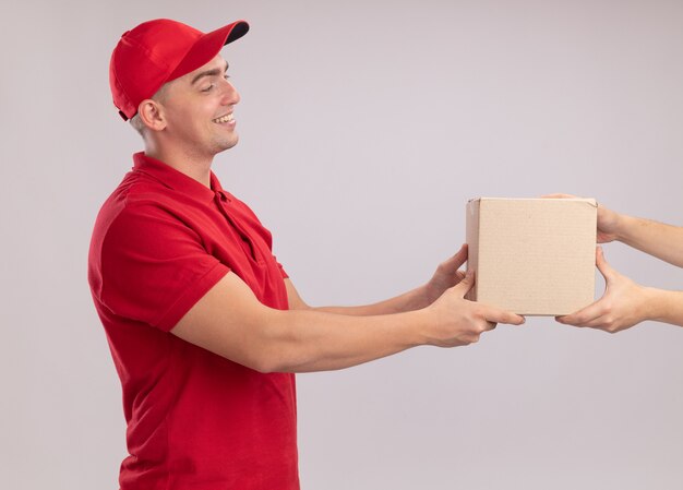 Smiling young delivery man wearing uniform with cap giving box to client isolated on white wall