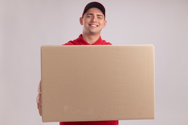 Улыбающийся молодой курьер в униформе с кепкой держит большую коробку, изолированную на белой стене