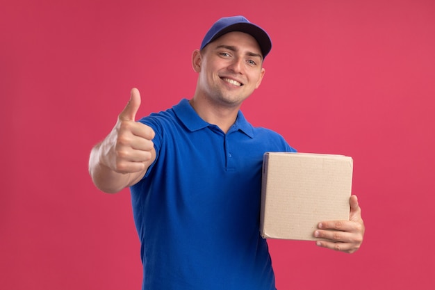 Улыбающийся молодой курьер в униформе с кепкой, держащей коробку, показывающую большой палец вверх, изолированную на розовой стене