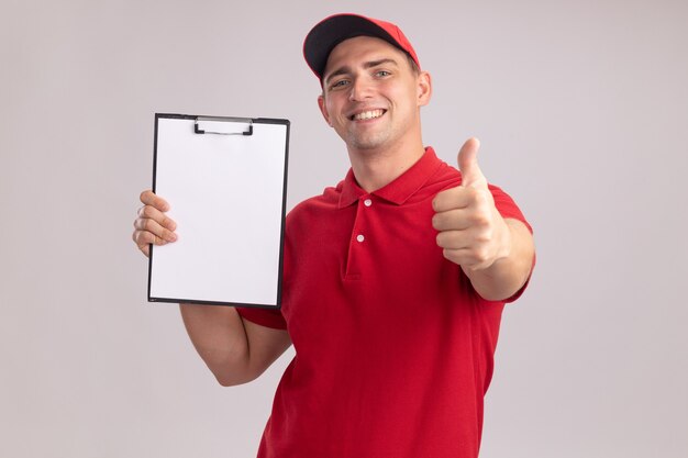 Smiling young delivery man wearing uniform with cap holding clipboard showing thumb up isolated on white wall