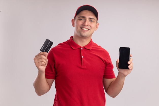 Smiling young delivery man wearing uniform with cap holding credit card with phone isolated on white wall