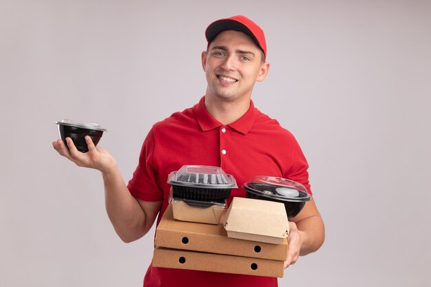 Smiling young delivery man wearing uniform with cap holding food containers on pizza boxes isolated on white wall