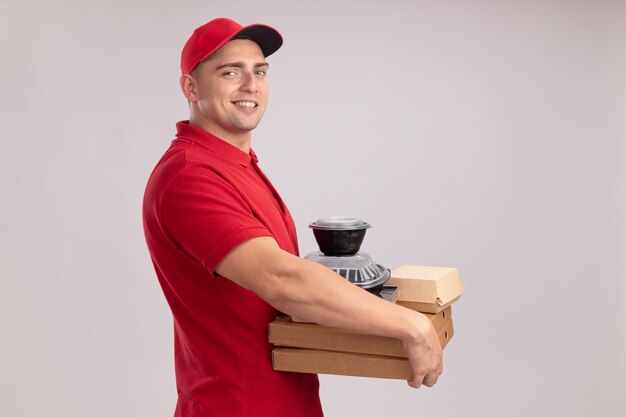 白い壁に隔離されたピザの箱に食品容器を保持するキャップと制服を着て笑顔の若い配達人