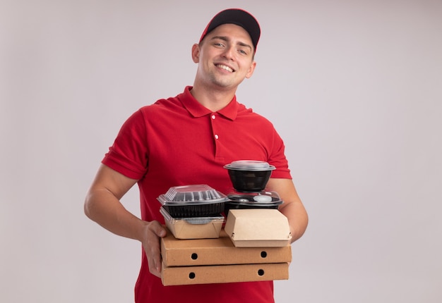 Smiling young delivery man wearing uniform with cap holding food containers on pizza boxes isolated on white wall