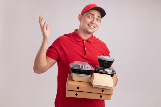 Smiling young delivery man wearing uniform with cap holding food containers on pizza boxes spreading hand isolated on white wall